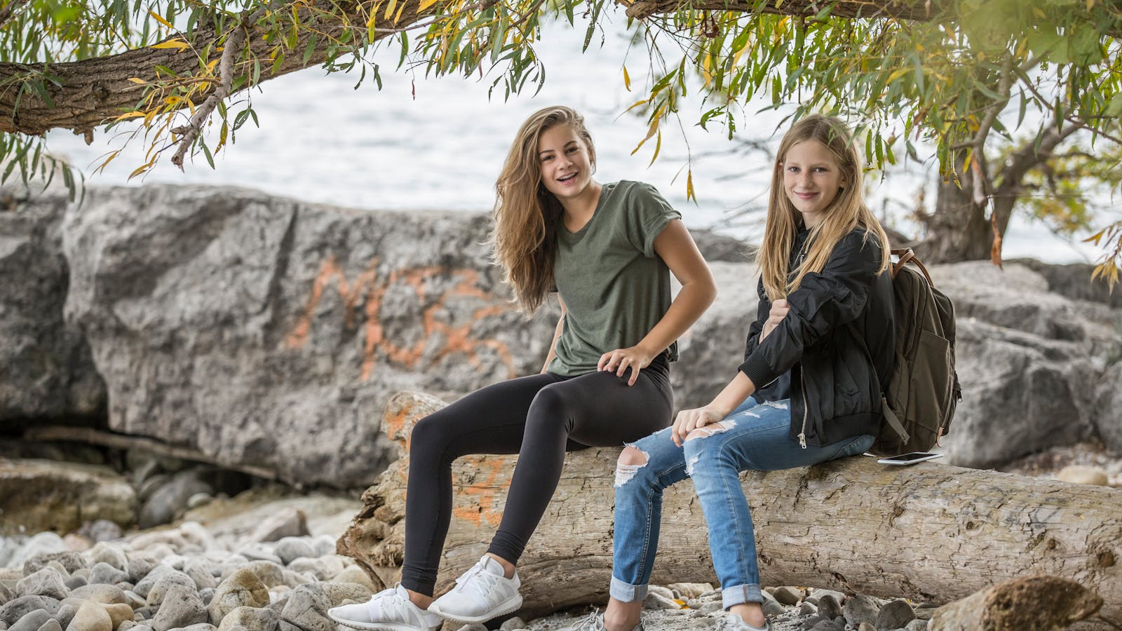 Two smiling teenage girls sitting on a log by the water, enjoying a casual day outdoors.
